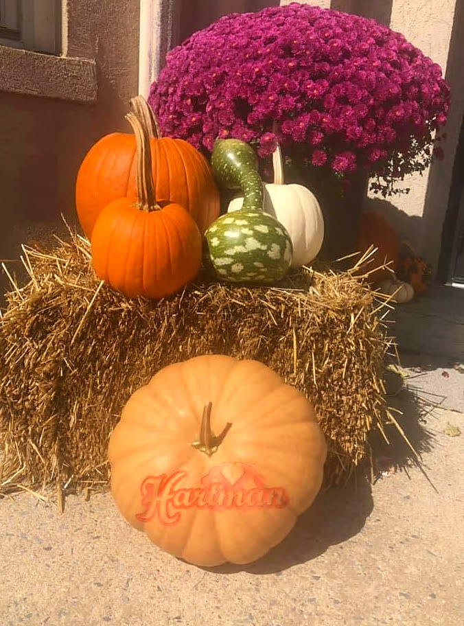 A porch display of hay bales, purple mums, and gourds with a pumpkin engraved Hartman in front