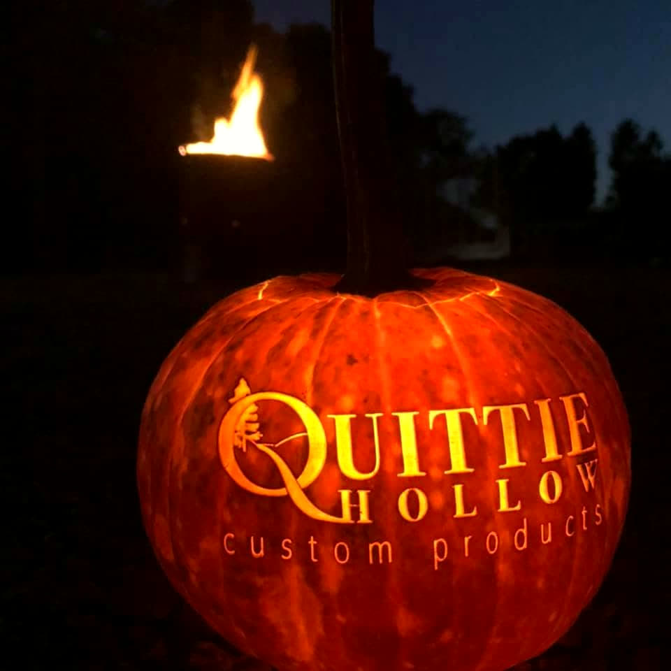 A pumpkin engraved with the Quittie Hollow logo, lit from the inside at dusk